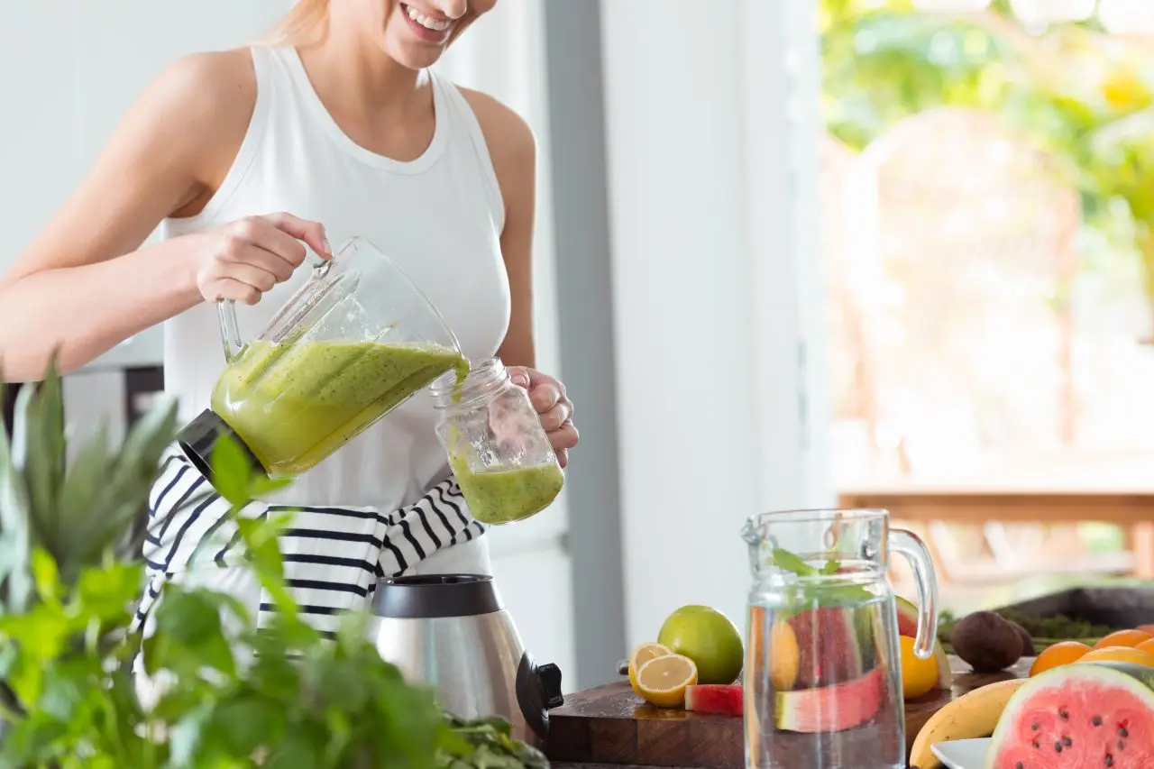 Happy woman on vegan diet, pouring green smoothie cocktail from mixer into glass in kitchen