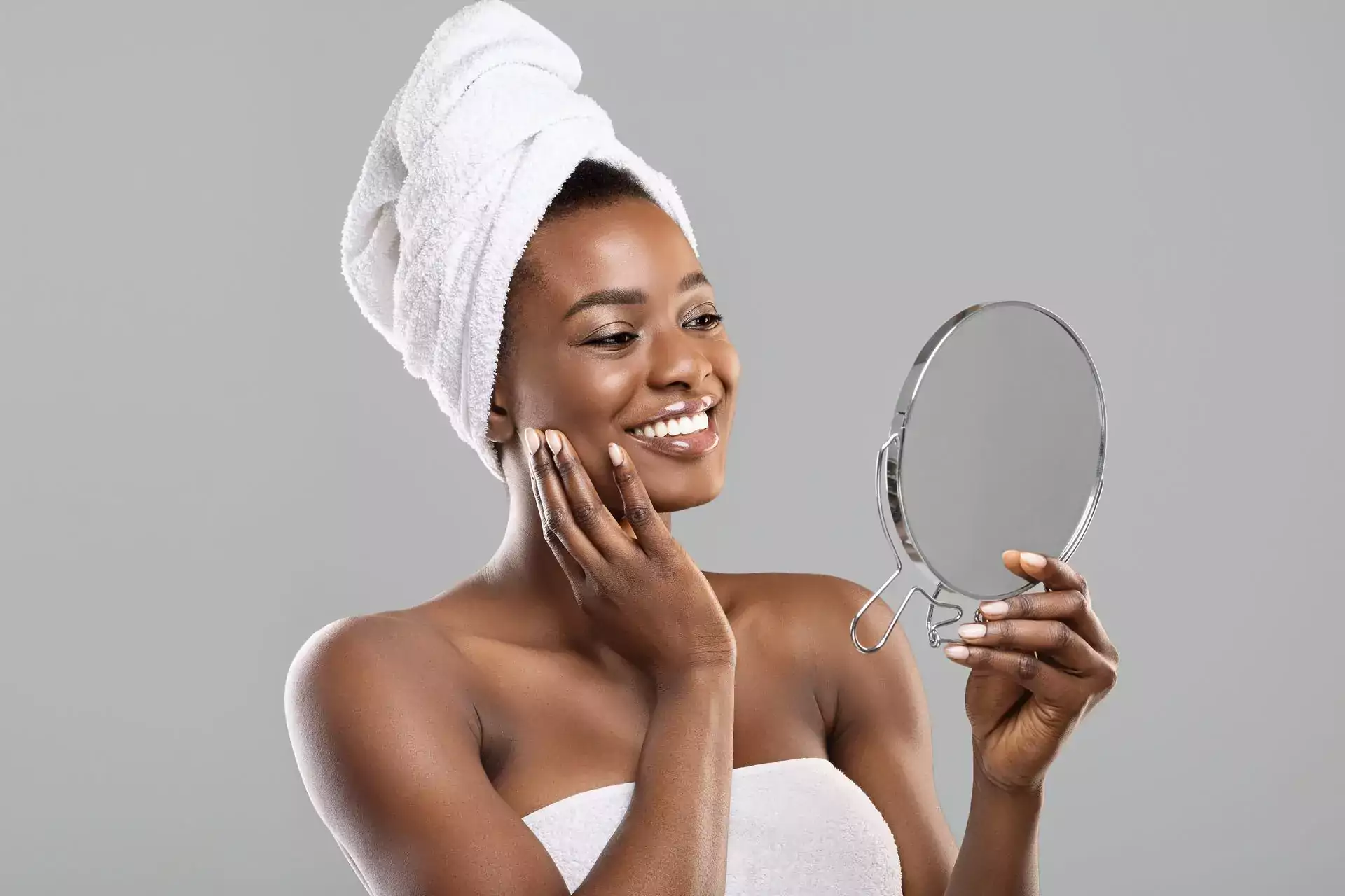 Beautiful afro woman in towel on head holding mirror and touching her perfect soft skin
