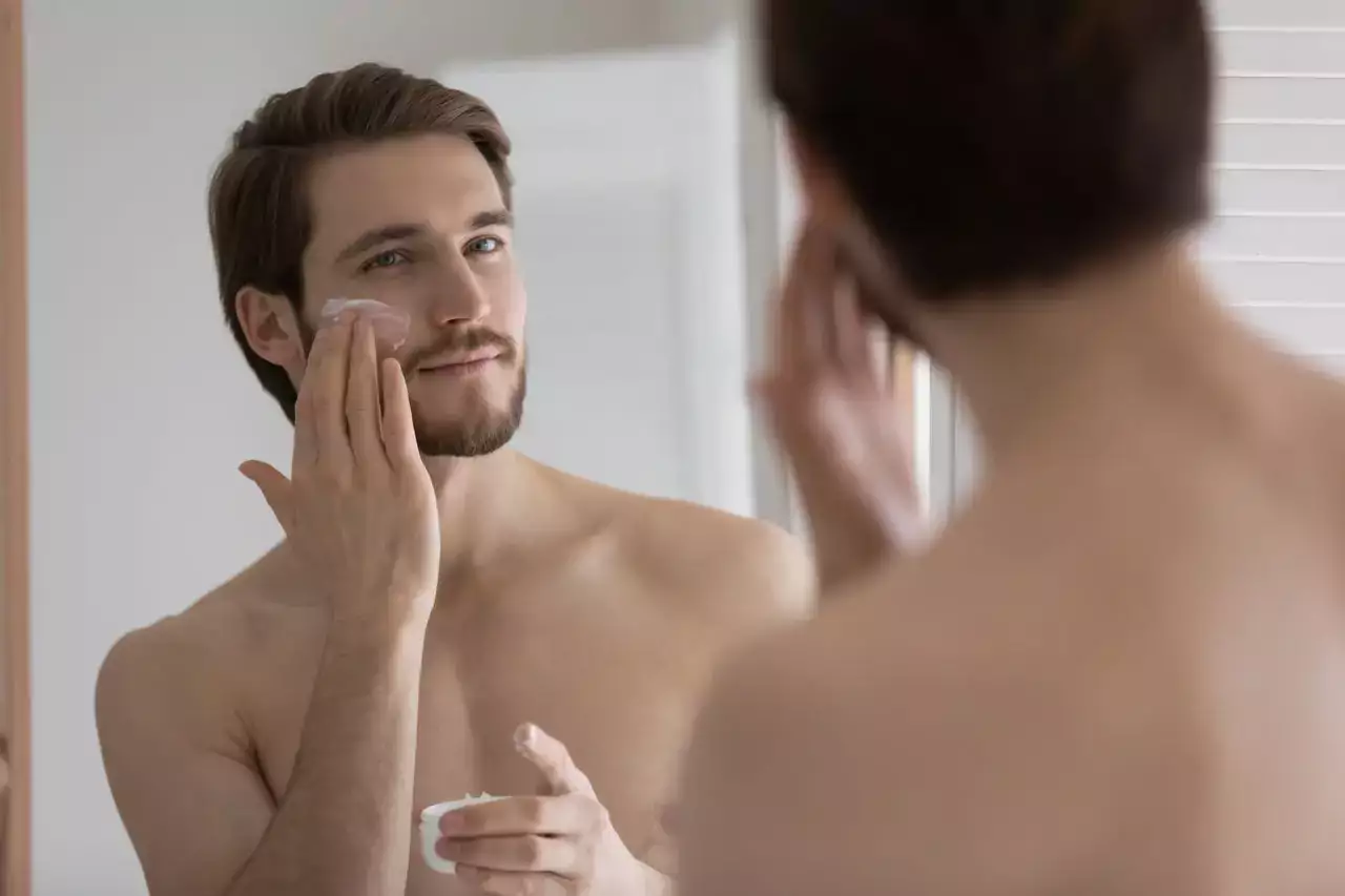 Handsome millennial man look in bathroom mirror after shower apply nourishing facial cream or mask
